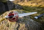 Pierced steel basket hilt broadsword seen from the pommel end, red liner showing through the bars, black scabbard behind on the rock