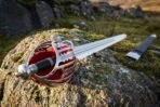 Close up of the pierced steel basket hilt with red cloth liner and black grip, broadsword resting on a mossy rock