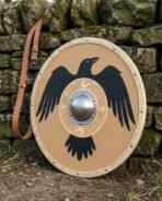 Planked round shield painted tan with a black raven, steel dome boss on an engraved knotwork roundel and rawhide rim leaning on a dry stone wall
