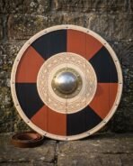Front view of a planked round shield in red and black quarters, riveted brass boss, knotwork ring and pale rim, coiled leather strap on the ground