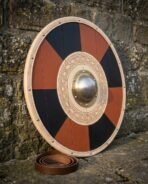 Planked round shield in red and black quarters with a brass boss on a carved knotwork ring and pale rawhide rim leaning on a stone wall