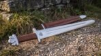 Steel Viking sword with a lobed pommel and tan leather grip laid on gravel beside its brown leather scabbard and a grassy bank