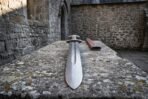 Point view of a Damascus steel Viking sword with a wide fuller and lobed pommel lying on a lichen covered stone slab