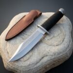 Polished 1095 steel sawback bowie knife with a clip point blade, sawtooth spine and black cord wrapped grip resting on a pale river stone beside a brown leather sheath