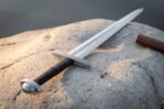 Polished arming sword with knotwork engraved crossguard, black leather grip and domed pommel on a pale sandstone rock