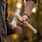 Polished bearded hand axe with struck rune marks and pale ash handle held in one hand in a sunlit autumn wood