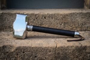 Polished blacksmith hammer with a Mjolnir shaped steel head, knotwork collar and black wrapped grip lying on a weathered stone step