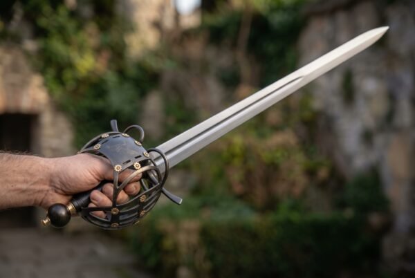 Polished broadsword with blackened basket guard and brass rosette rivets held out in a fist in front of an overgrown ruin