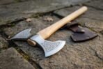 Polished double bit axe head with Celtic knot etching and light ash haft lying on stone slabs with its cover