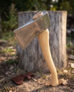 Polished felling axe with knotwork medallion on the cheek and curved ash handle leaning on a chopping stump, leather cover below