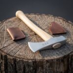 Polished hand axe head with rune marks and wooden wedge seen from the handle end on a stump with leather sheath and pouch