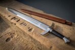 Polished hand forged viking short sword with a triple fullered blade and a downswept crossguard resting on a weathered sandstone block beside a brown leather scabbard