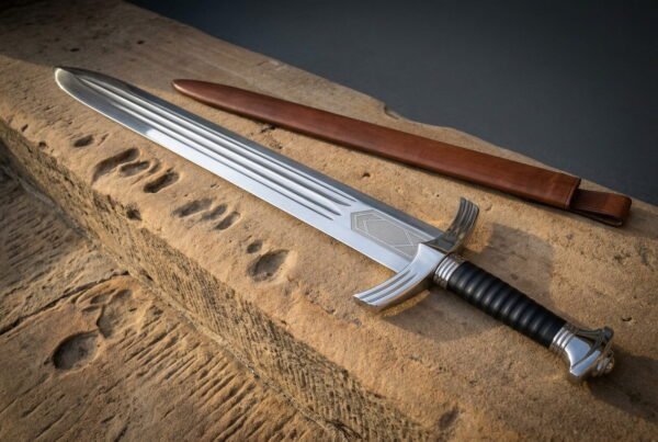 Polished hand forged viking short sword with a triple fullered blade and a downswept crossguard resting on a weathered sandstone block beside a brown leather scabbard