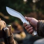Polished kukri knife with etched knotwork band and stag horn handle held in a hand beside a leather sheath with companion blades