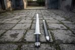 Polished Norse sword with broad fuller, black grip and lobed steel pommel lying on cobbles next to its black scabbard