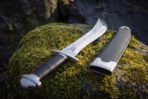 Polished sawback short sword and black leather sheath lying on a mossy woodland boulder, showing the stacked leather grip, domed oval steel pommel and saw toothed spine.