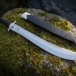 Polished sawback short sword with an upswept clip point and black leather grip resting on a moss covered boulder beside a black scabbard with steel throat and chape