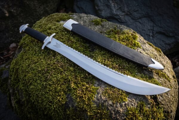 Polished sawback short sword with an upswept clip point and black leather grip resting on a moss covered boulder beside a black scabbard with steel throat and chape