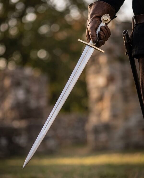 Polished steel arming sword with brass wheel pommel held point down in a gloved hand in front of blurred ruins