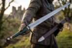 Polished steel arming sword with green leather grip and blackened quatrefoil guard held out by a man before castle ruins