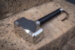 Polished steel blacksmith hammer with Mjolnir head, steel end cap and leather wrist loop resting on sandstone, side view