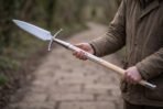 Polished steel boar spear with broad leaf blade, curved crossbar and riveted socket on a pale ash haft held by a man on a path
