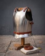Polished steel breastplate on a wooden stand, angled to show the roped edge, red leather shoulder straps and waist belt, folded tunic beneath