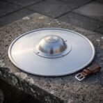Polished steel buckler with Mjolnir engraved domed boss and rolled rim lying on a granite block beside a leather strap