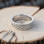 Polished steel Celtic knotwork ring standing on a pale sawn log showing the runes cut inside the band, a suede pouch in front