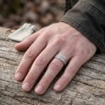 Polished steel Celtic knotwork ring worn on a man's finger, hand resting on weathered wood beside a small suede pouch