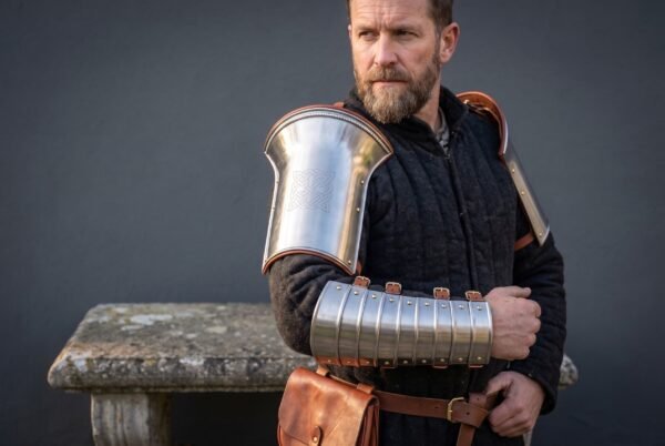 Polished steel flared bracer on the upper arm and articulated manica on the forearm worn by a bearded man in a dark quilted jacket