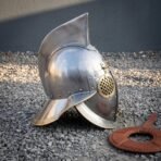 Polished steel gladiator helmet in side view showing the wide brass edged brim and crest fin on gravel
