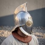 Polished steel gladiator helmet worn by a man in a linen shirt, brass grille eye disc and leather collar in view
