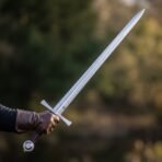 Polished steel knightly arming sword with steel wheel pommel and straight cross held aloft in a gloved hand in the woods