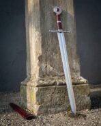 Polished steel knightly sword with oxblood grip standing against a stone pillar, red scabbard lying on the gravel