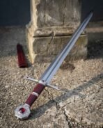 Polished steel knightly sword with red stone octagonal pommel and oxblood grip lying on gravel by its red scabbard
