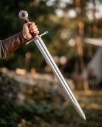 Polished steel knightly sword with red stone pommel and set guard held point down in a gloved hand among trees