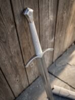Polished steel longsword, close up of the shield pommel, cream cord wrapped grip and arched guard by a wooden wall