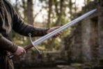 Polished steel longsword held in both hands near a ruined stone wall, curved cross guard and leather grip in view