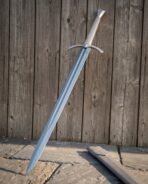 Polished steel longsword leaning at an angle on wooden boards over stone paving with its pale scabbard below