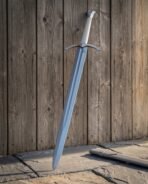 Polished steel longsword resting against a weathered plank wall in warm light, pale grey scabbard on the flagstones