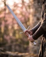 Polished steel longsword with arched crossguard held in two hands against blurred pink blossom