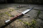 Polished steel longsword with two hand leather grip and steel pommel lying diagonally on mossy paving with its scabbard behind