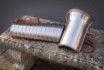 Polished steel manica arm guard of articulated bands laid beside a flared bracer with etched knotwork and russet leather edging on a stone bench