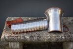 Polished steel manica arm guard of articulated bands beside a flared knotwork engraved bracer with russet leather straps on a stone bench outdoors.