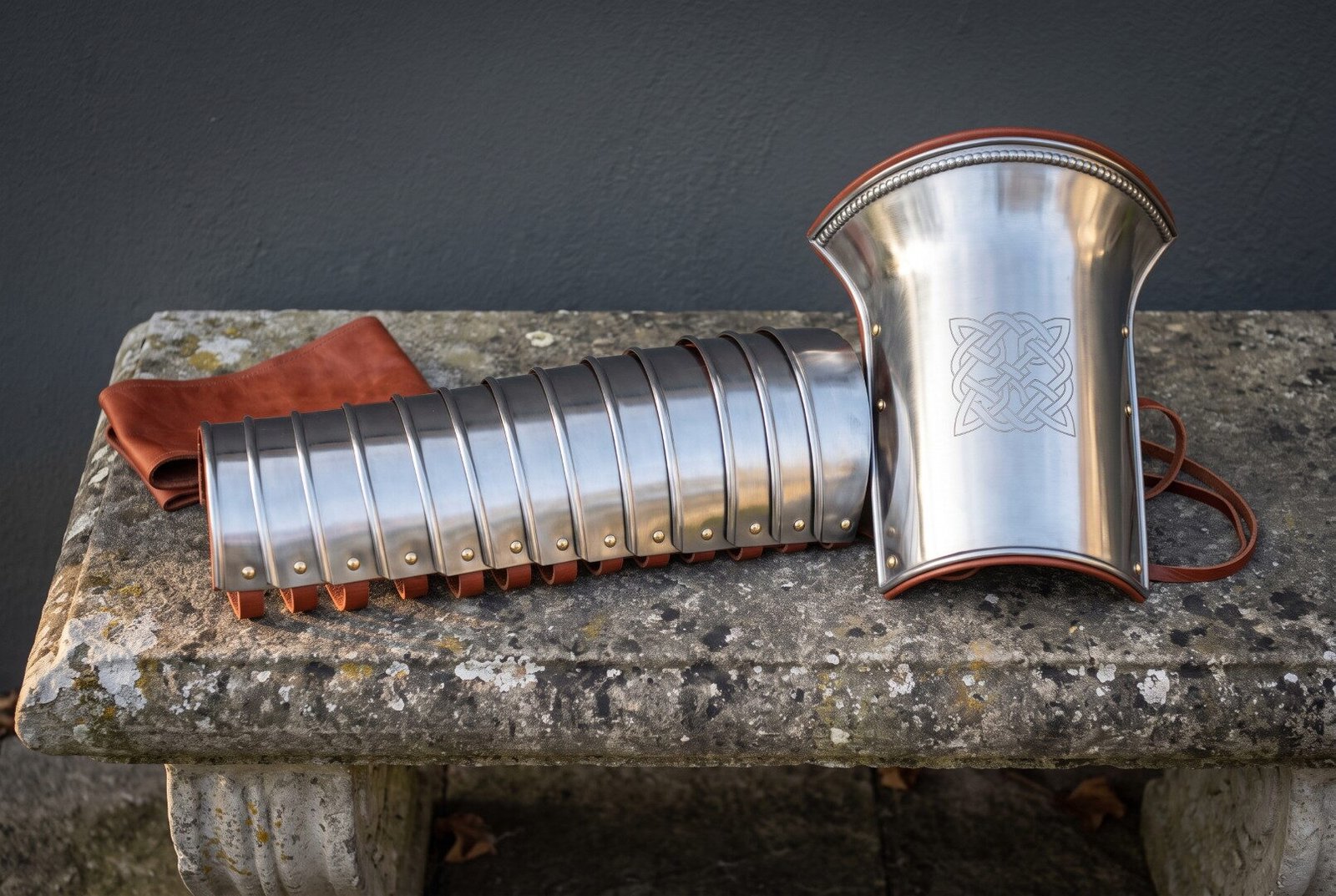 Polished steel manica arm guard of articulated bands beside a flared knotwork engraved bracer with russet leather straps Polished steel manica arm guard of articulated bands beside a flared knotwork engraved bracer with russet leather straps on a stone bench outdoors.