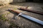 Polished steel medieval longsword with pierced quatrefoil guard and leather grip lying on stone flags beside its brown scabbard