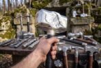Polished steel Mjolnir style hammer with wrapped black grip held up in a hand above a table of forged hammers outdoors