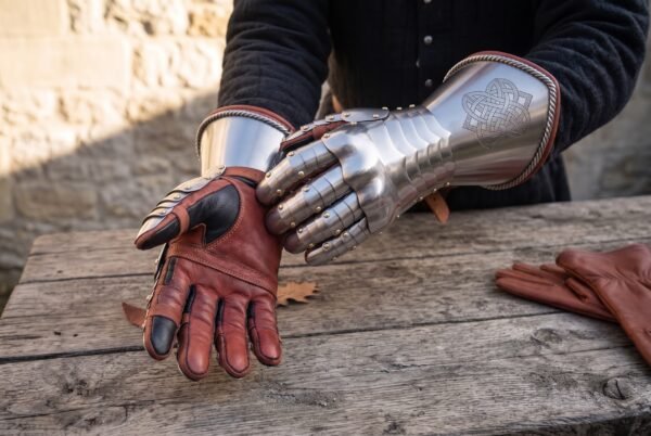 Polished steel plate gauntlet with a knotwork etched cuff being pulled onto a red leather glove at a wooden table