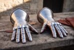 Pair of polished steel plate gauntlets with articulated finger lames and knotwork etched cuffs laid on a wooden table with leather gloves